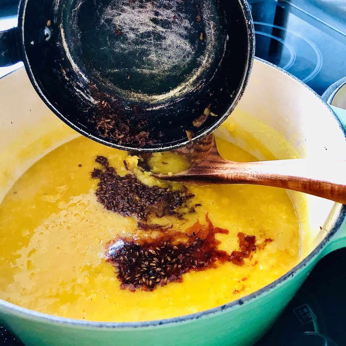 Pouring tadka from a small frying pan into a large heavy pot of cook daal.