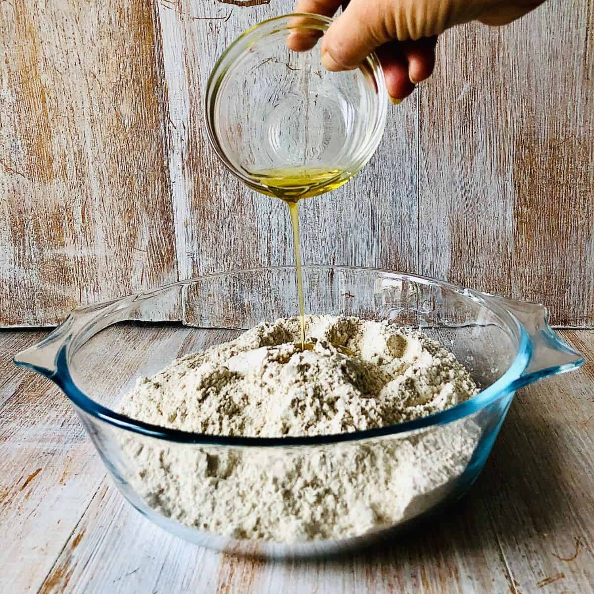 A glass bowl containing chapati flour, with olive oil being poured into the flour from a small glass dish.