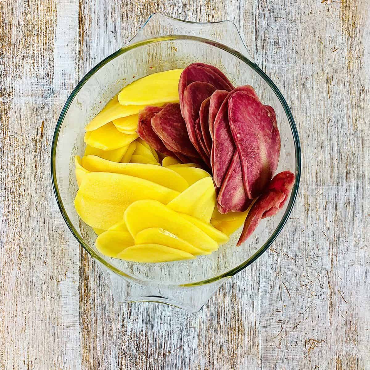 A glass bowl containing thin slices of Yukon gold and Heidi red potatoes coating with melted butter and rose harissa.