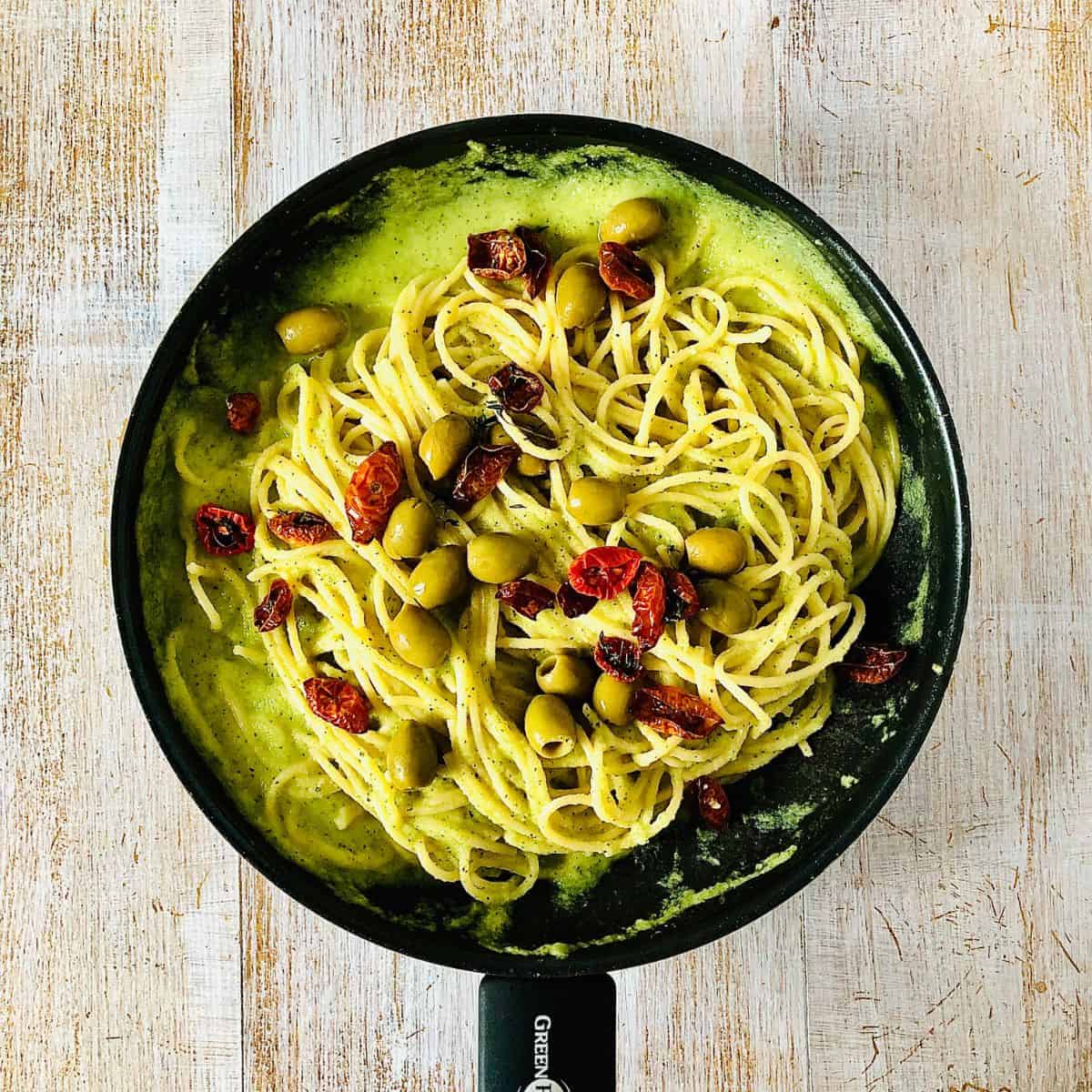 A frying pan containing spaghetti pasta in a courgette sauce with dried cherry tomatoes and olives.