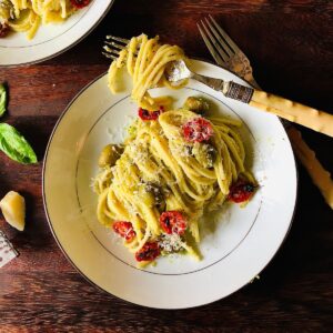 A plate of spaghetti pasta in a courgette sauce topped with dried cherry tomatoes, green olives and parmesan cheese.