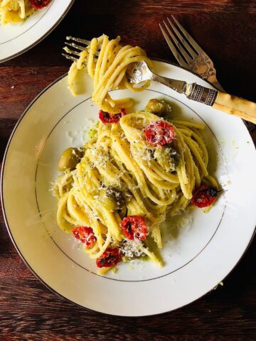 A plate of spaghetti pasta in a courgette sauce topped with dried cherry tomatoes, green olives and parmesan cheese.