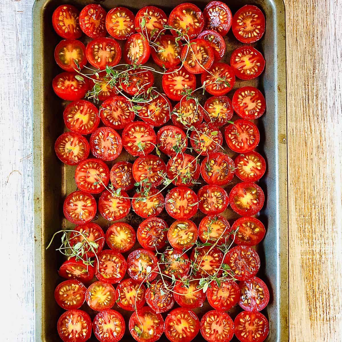 A baking tray filled with cherry tomatoes sliced in half with sprigs of thyme, ready for cooking.