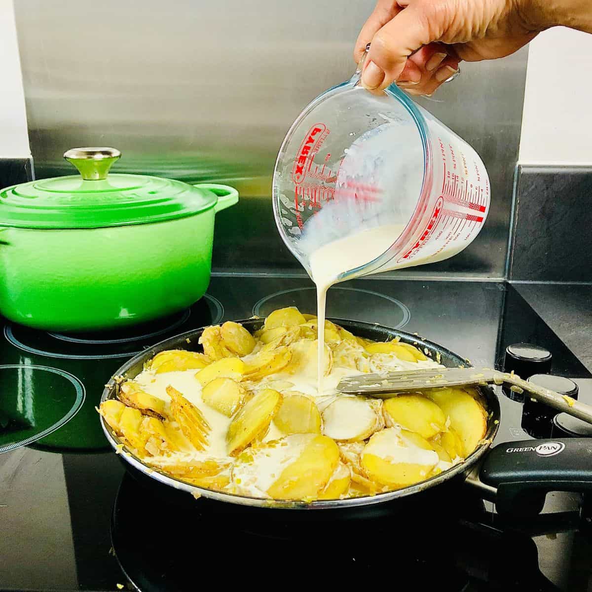Pouring vegan cream from a measuring jug into a frying pan on a hob containing thinly sliced potato.