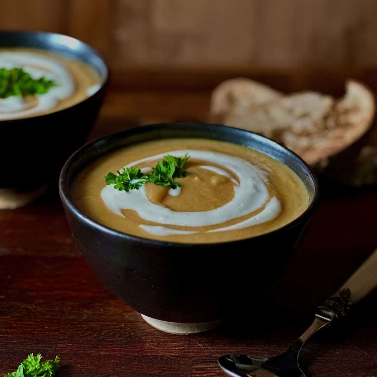 A dark brown serving bowl containing winter vegetable soup with a vegan yoghurt garnish and a sprig of parsley.