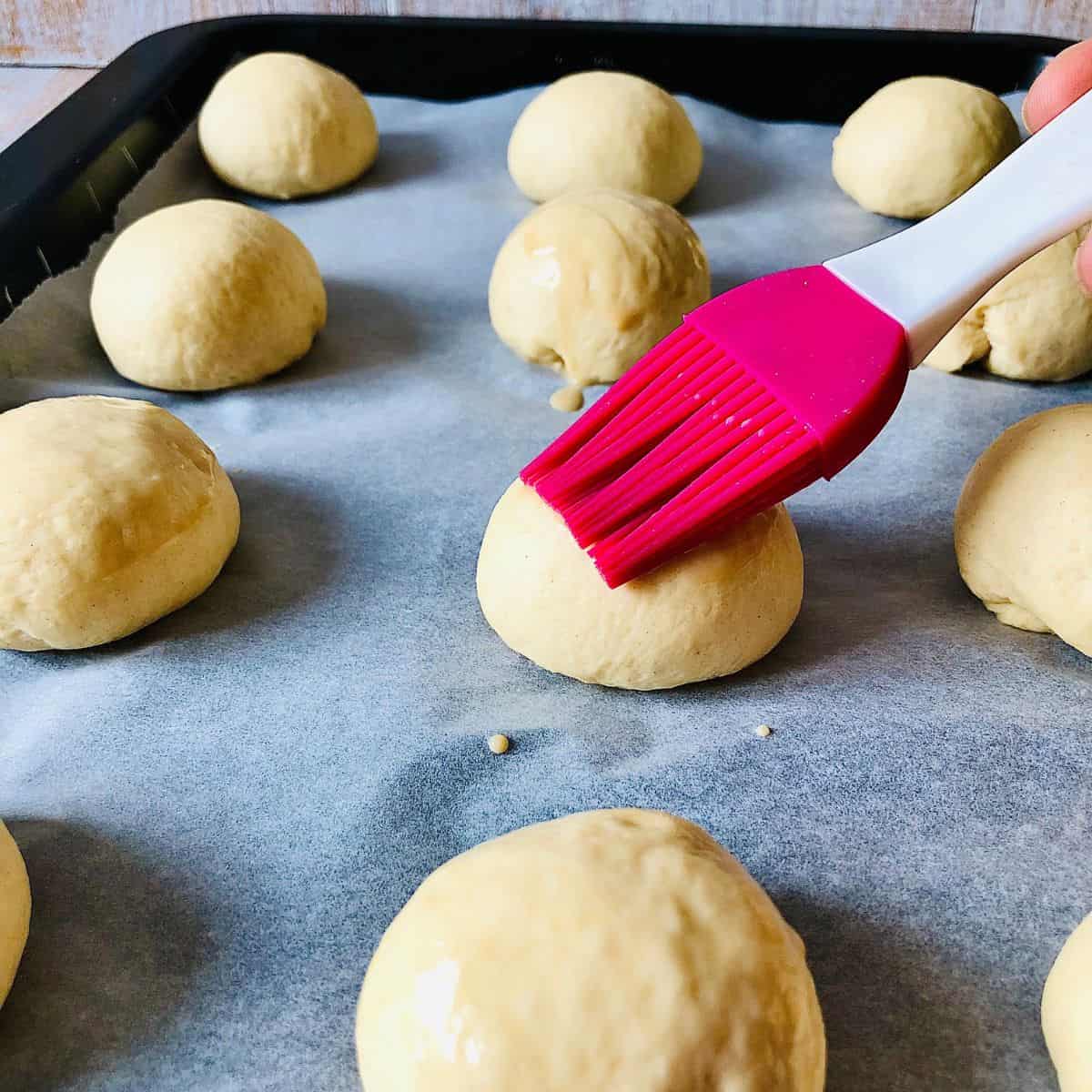 A hand Brushing mini brioche bun dough balls with plant-based milk/soy sauce.