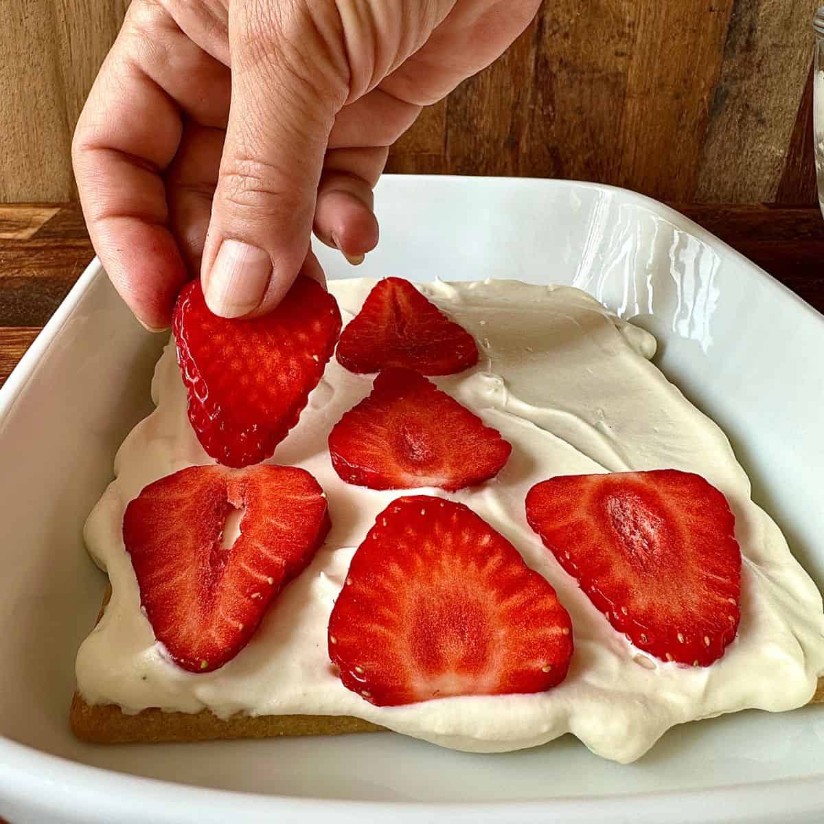 A white deep dish containing the first layer of an elderflower and strawberry cake. A layer of biscuit topped with a layer of whipped cream and thinly sliced strawberries.