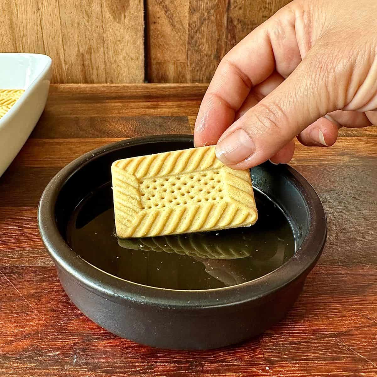 A hand placing a biscuit into a shallow dish containing elderflower cordial for soaking.