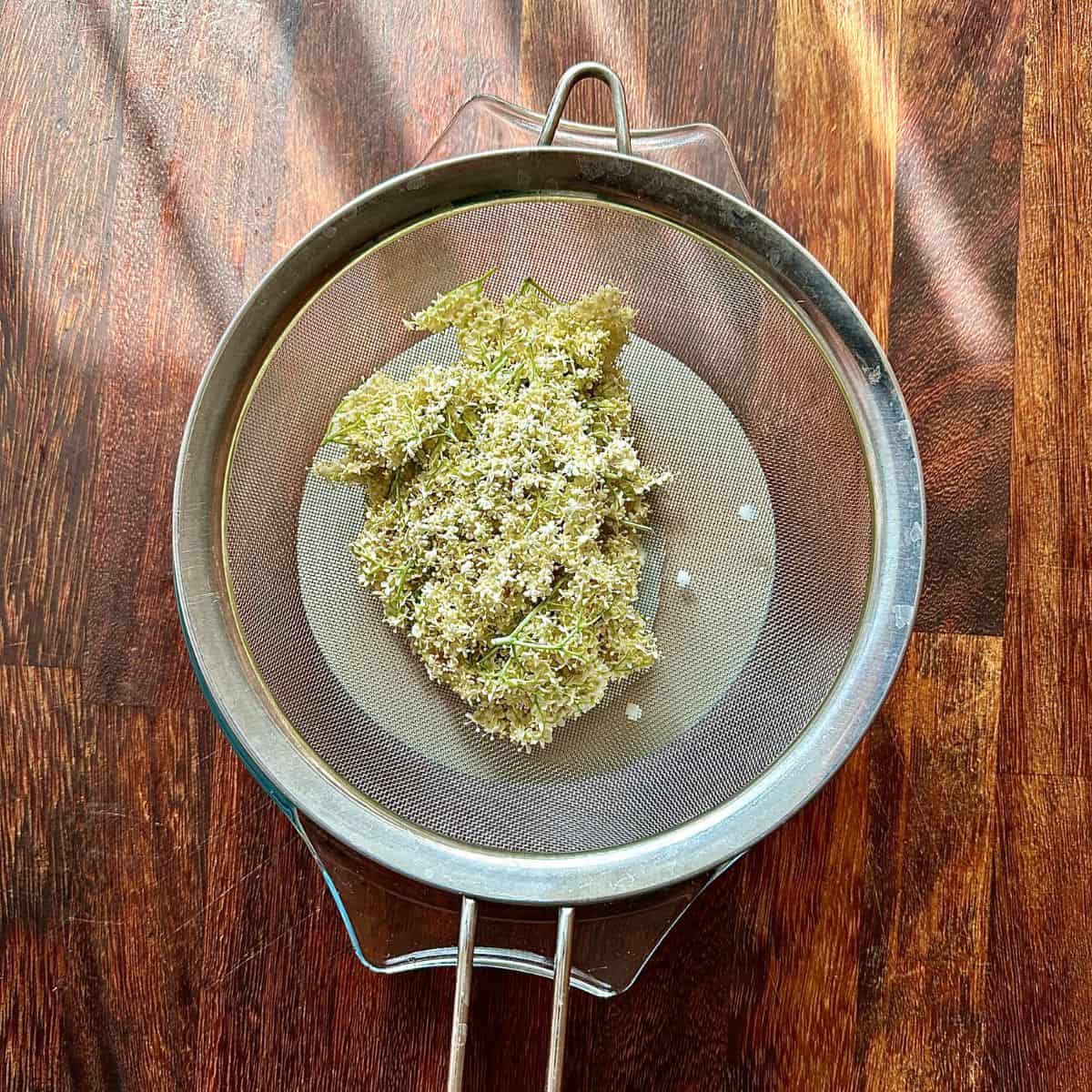 Elderflower heads in a sieve over a glass bowl containing strained infused vegan cream.