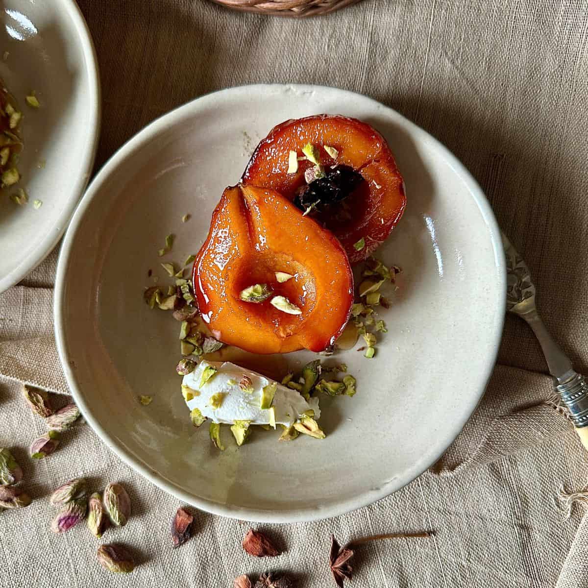 Two halves of baked quince with a liquor glaze, cream cheese quenelle, and chopped pistachio.In a beige serving bowl.