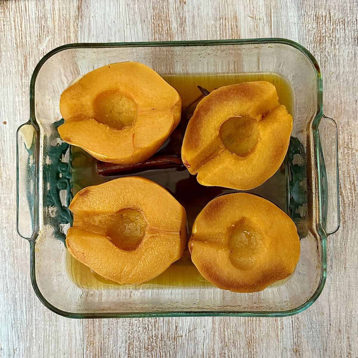A glass oven dish containing four quince halves, sitting in a liquor,, after baking, cut side up.