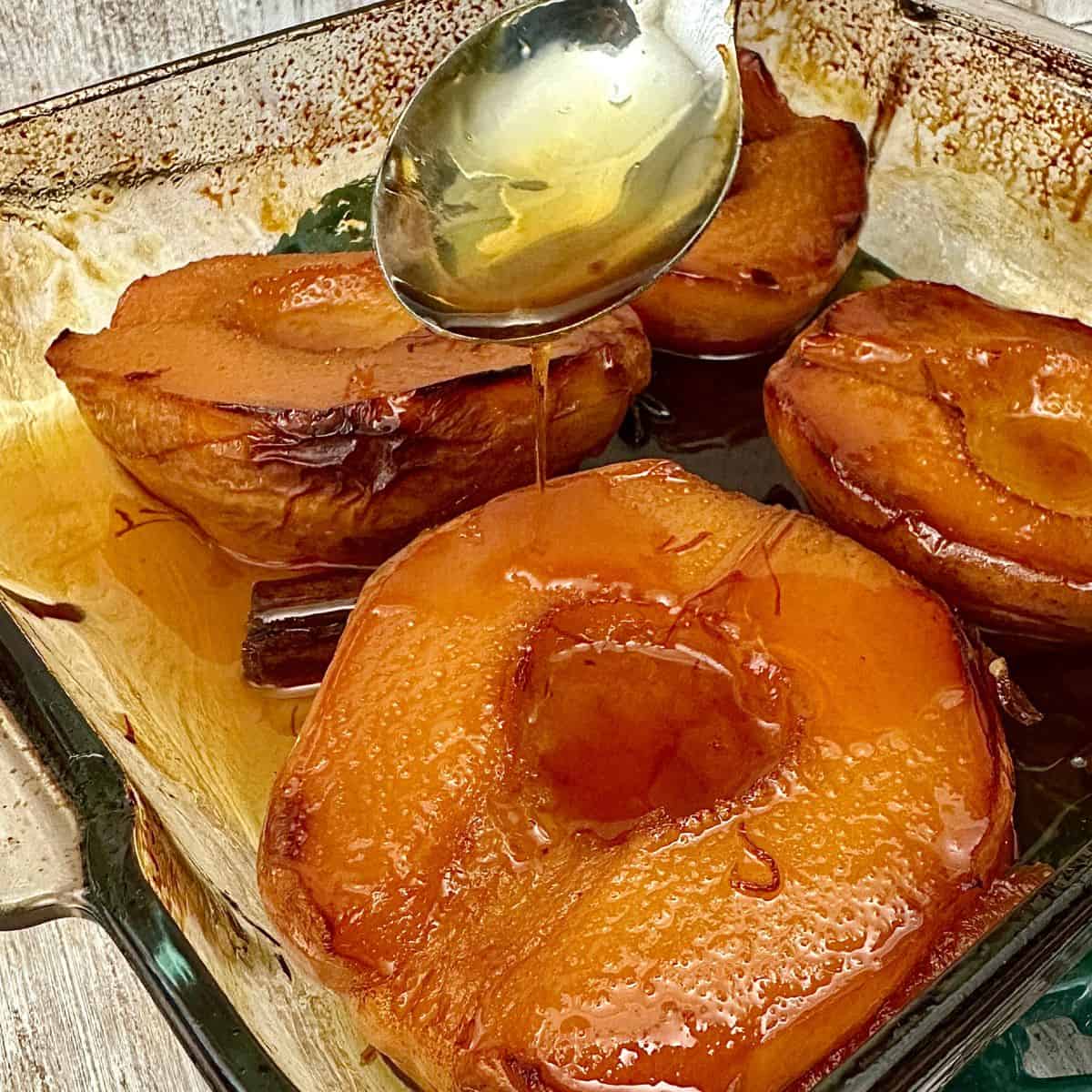 Four baked quince halves covered with a liquor glaze in a glass oven dish. The liquor is pouring from a dessert spoon onto the quince.