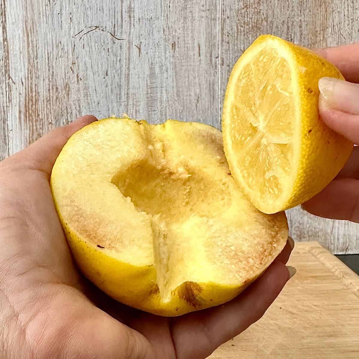 Close up of half a quince fruit held in a hand with the other hand holding half a lemon ready to rub the lemon flesh on the quince flesh to prevent oxidation.
