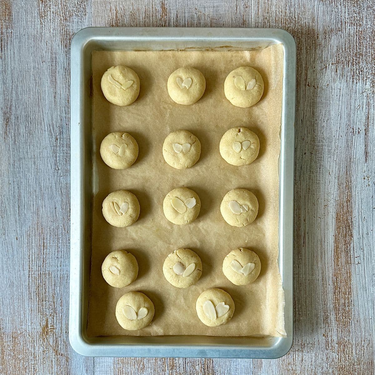 Fourteen Gulab Jamun Cookie dough balls topped with slivers of almond on a baking tray lined with parchment paper, ready to bake.