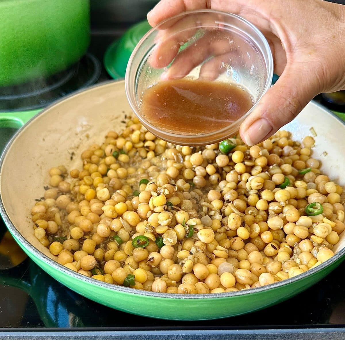 A hand pouring tamarind pulp from a small glass dish into a saucepan containing cooked yellow peas.