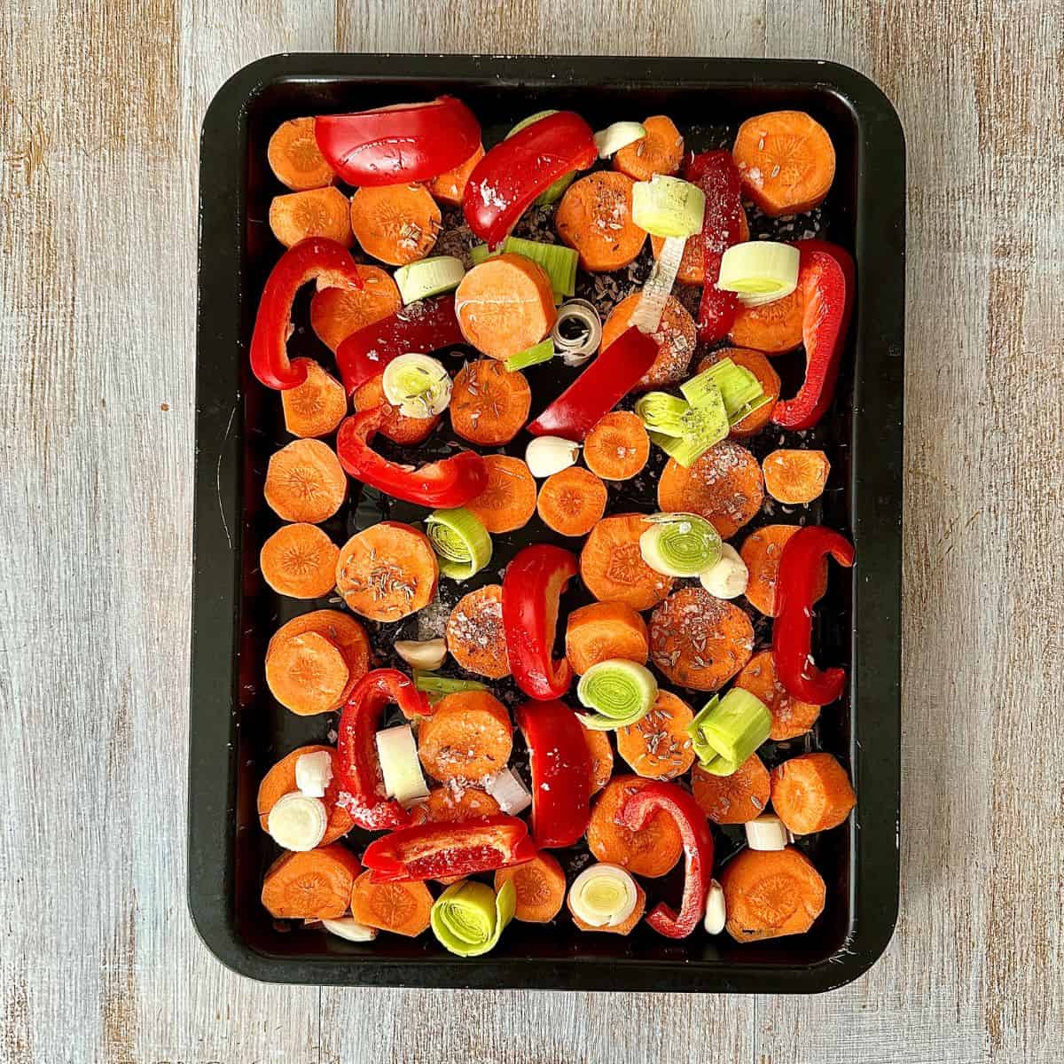 A sheet pan containing chopped carrot, leek, red peppers whole garlic and fennel seeds, ready for roasting.