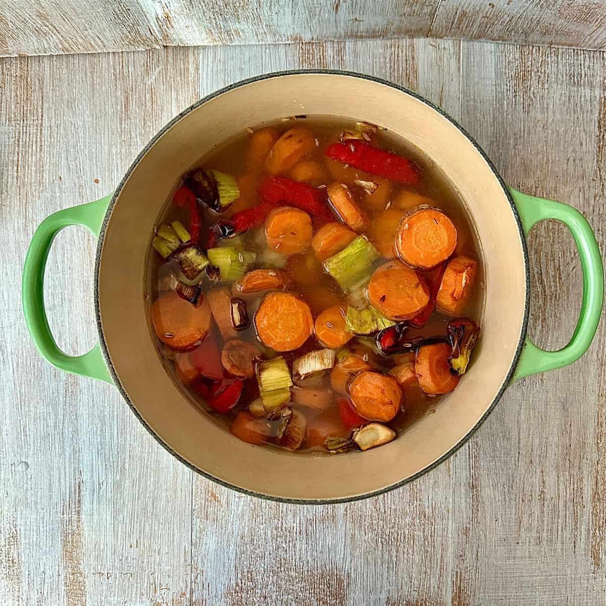 A large green pot with two side handles containing roasted chopped vegetables and vegetable stock for carrot and red pepper soup, ready to blend.