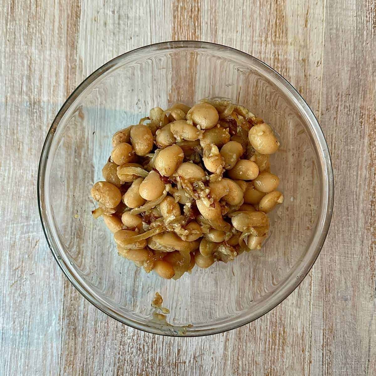 A round glass bowl containing cooked butterbeans with caramelised onion ready to puree.