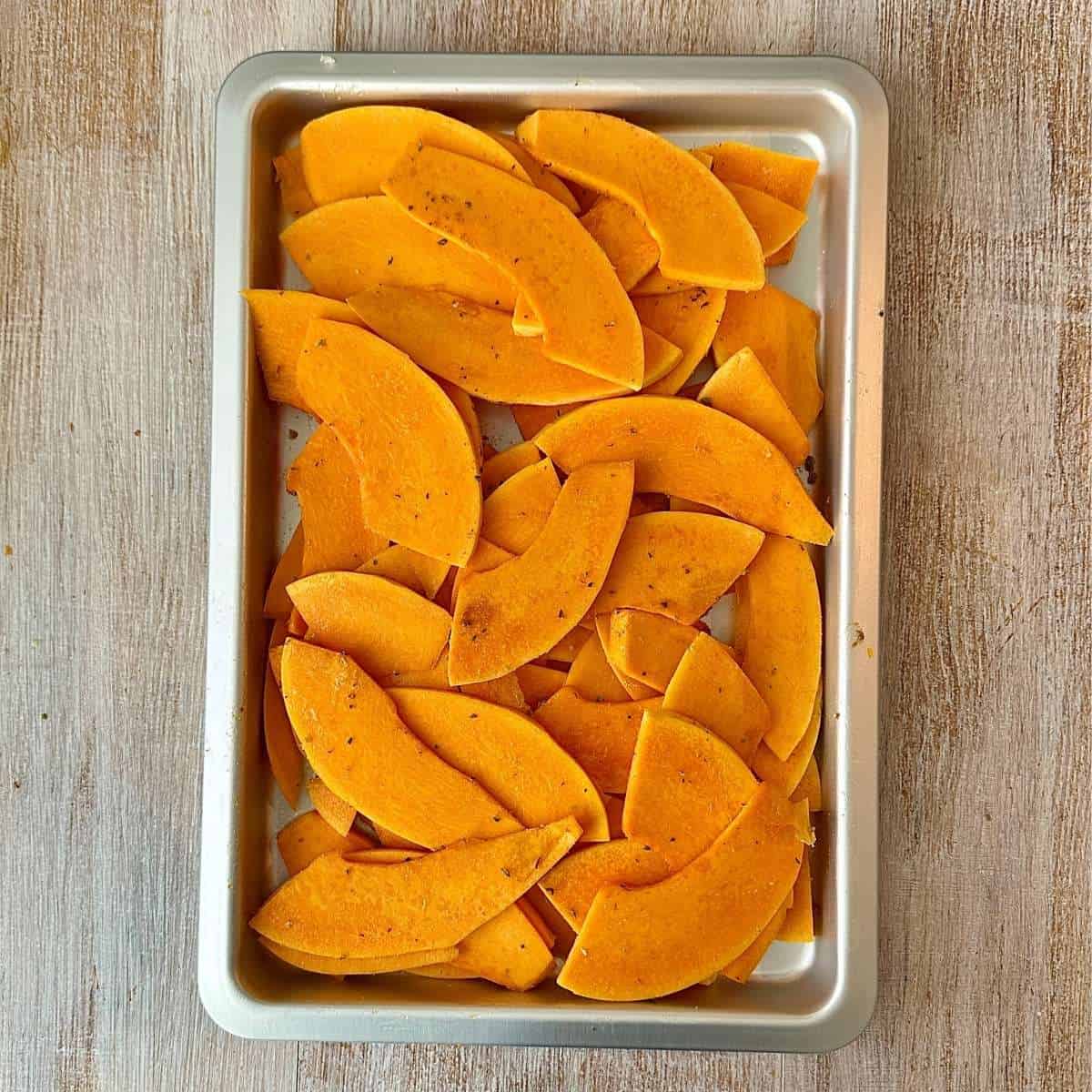 A baking tray containing slices of uncooked crown prince pumpkin seasoned with cumin and oregano.