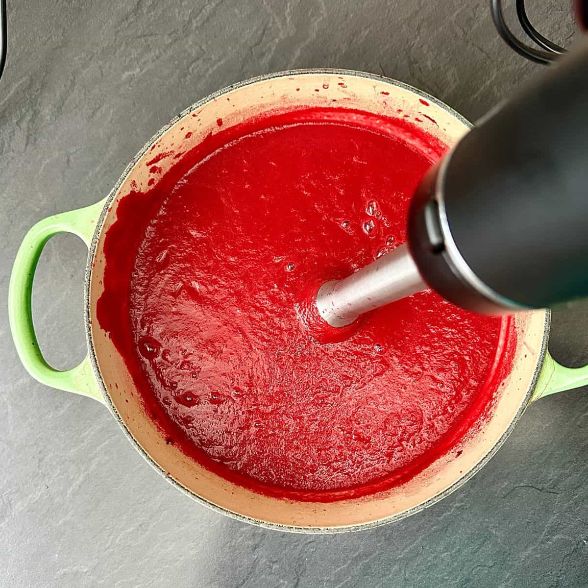 A large green pot containing blended beetroot soup. A stick blender Is being used to blend the soup.