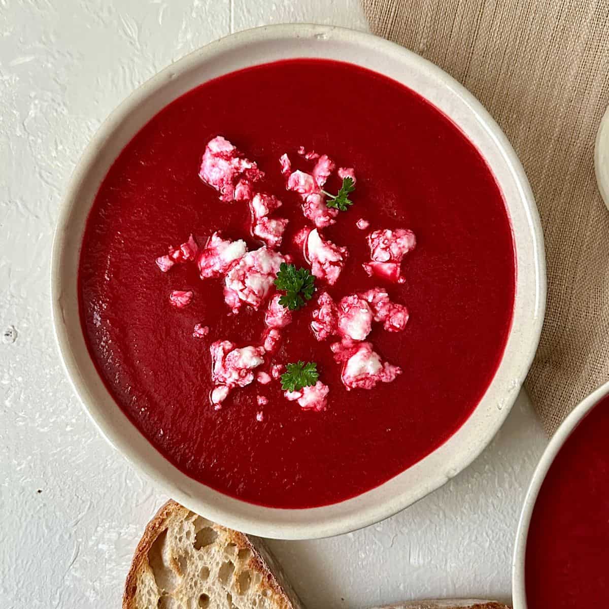 A beige coloured ceramic bowl containing beetroot soup garnished with vegan feta cheese and parsley.