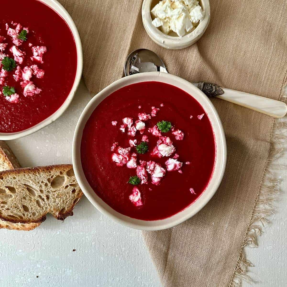 Two beige ceramic bowls containing beetroot soup garnished with vegan feta cheese.