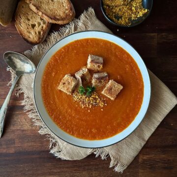 A white bowl containing carrot and red pepper soup with croutons.