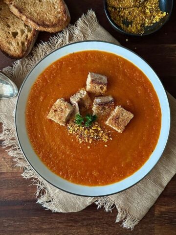 A white bowl containing carrot and red pepper soup with croutons.