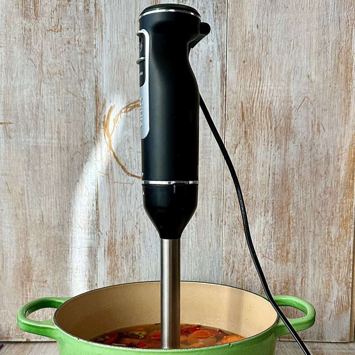 An electric stick blender stood upright in a large green pot containing chopped vegetables and stock for carrot and red pepper soup, ready to blend.
