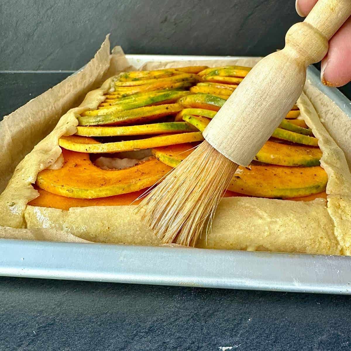 Close up of a brush coating the pastry of a pumpkin galette with an egg wash.