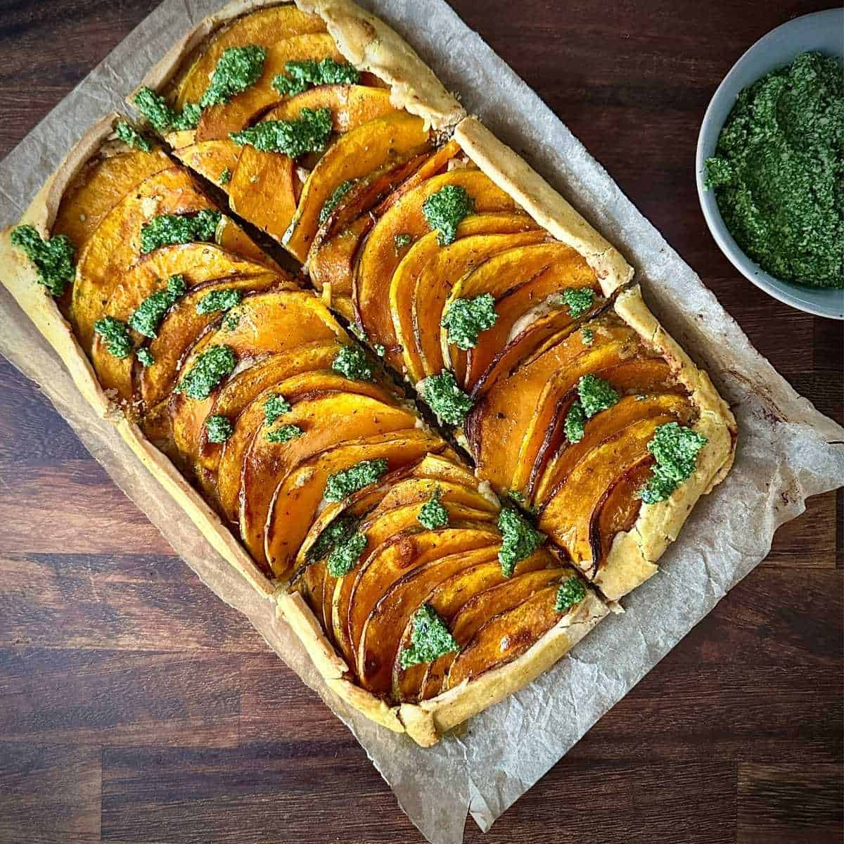 A cooked crown prince pumpkin galette with rocket pesto, cut into six portions, sitting on top of parchment paper with a bowl of rocket pesto next to it.