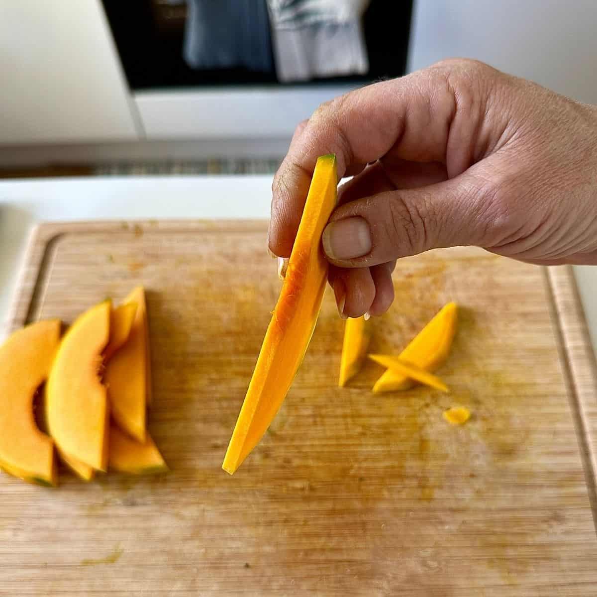 A hand holding a thin slice of uncooked crown prince pumpkin showing the thickness of the slice.