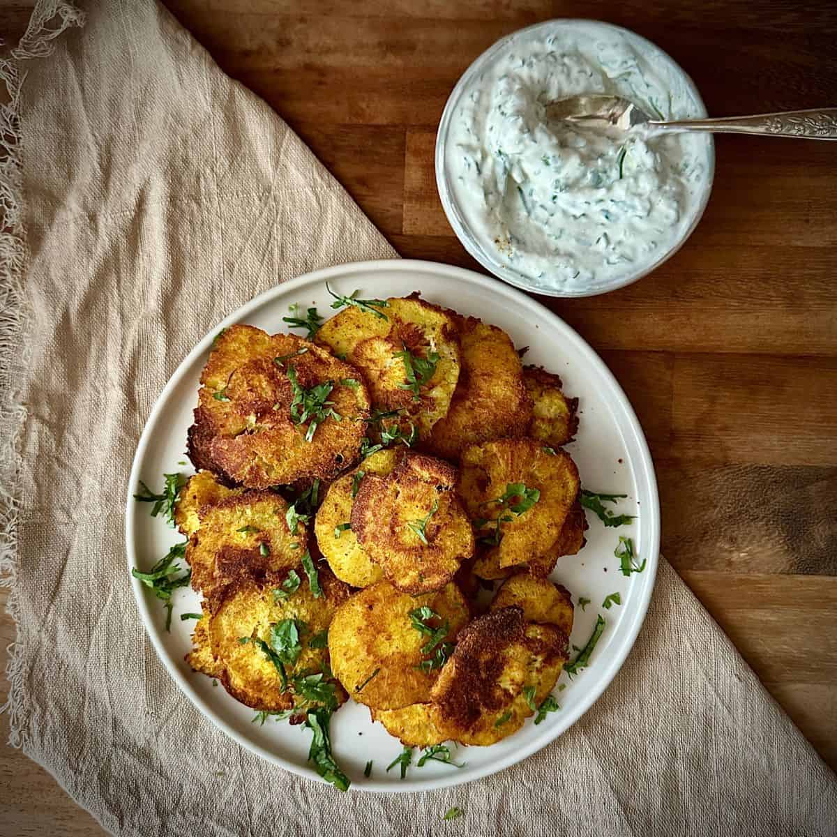 A white ceramic plate with a pile of smashed roast parsnips with a smaller dish containing a yoghurt tahini dip next to it.