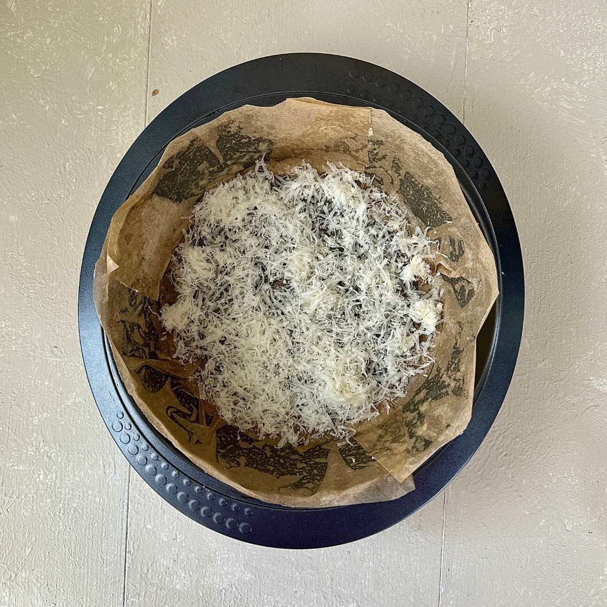 A round cake tin lined with parchment paper with a layer of grated vegan cheese on the bottom.