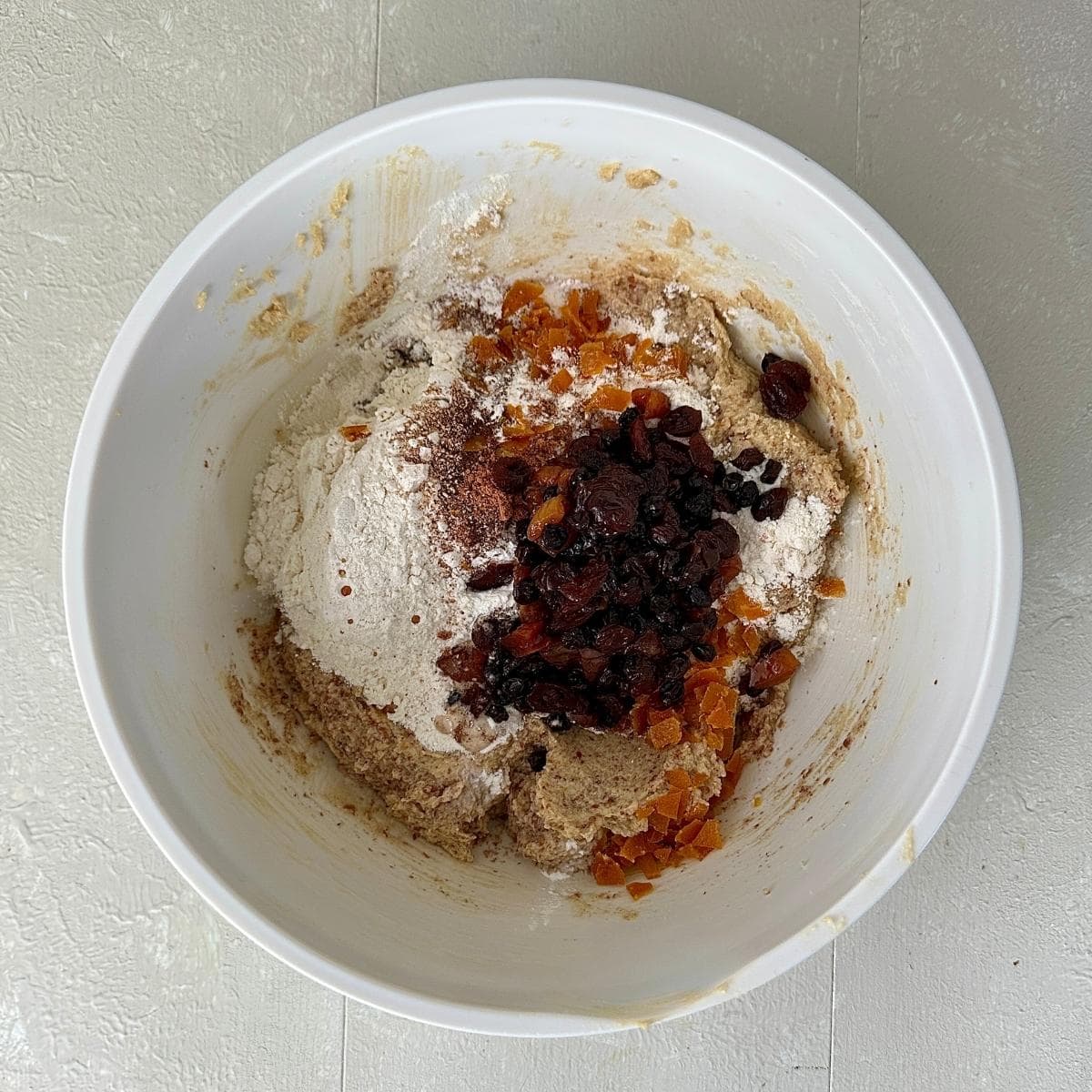 A white bowl containing mixture for vegan Christmas cupcakes. Dried fruit infused with alcohol, baking powder and candied orange pieces sits on top of creamed soft brown sugar, vegan butter, flax-egg and ground almond. All ready to be mixed together.