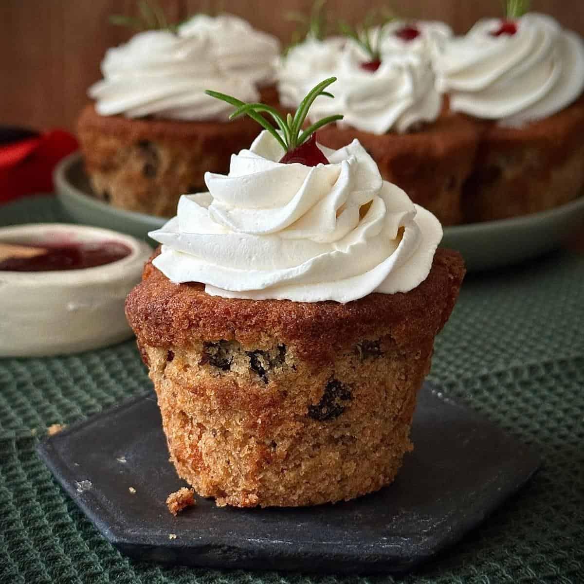 Close up of a vegan Christmas cupcake with brandy frosting piped on top and decorated with cranberry jam and a sprig of rosemary.