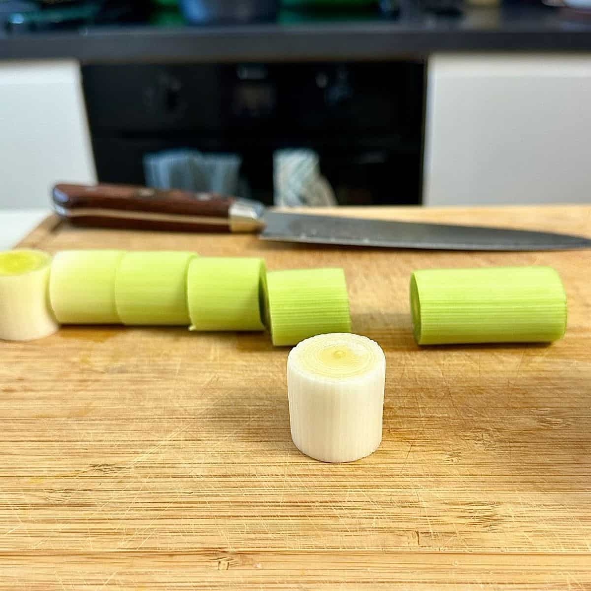 A leek chopped into three-quarter inch length on a wooden chopping board. One piece sits on its cut edge in the foreground.