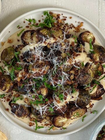 A white plate with crispy roasted Brussels sprouts in a vegan cheese sauce. Sprinkled with grated vegan parmesan and parsley.