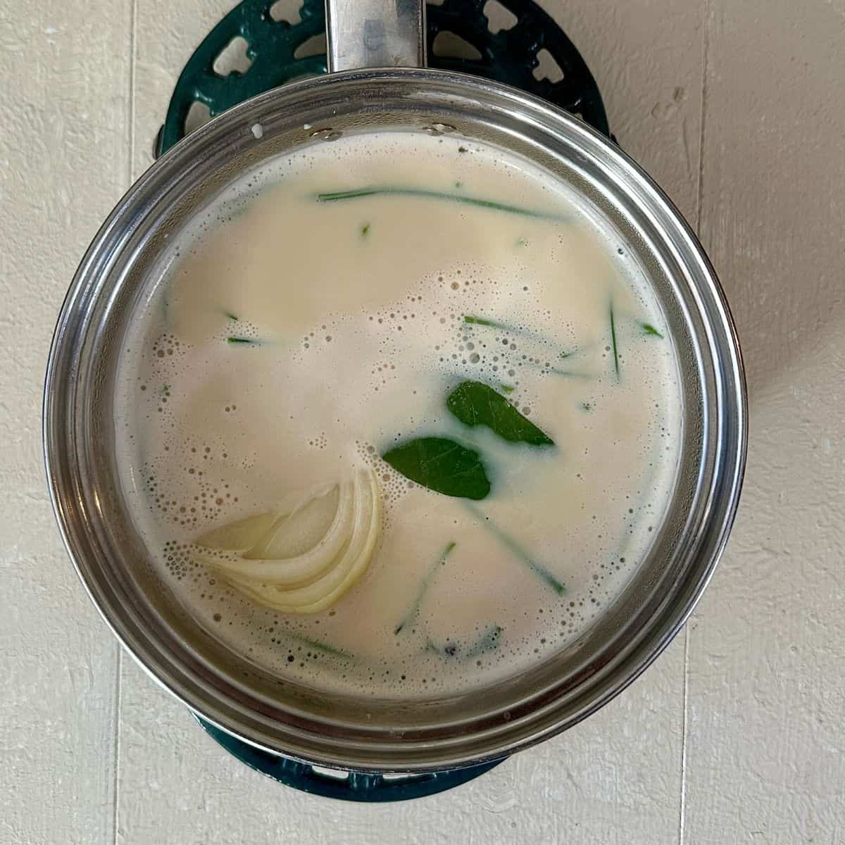 A steel saucepan containing plant-based milk with a wedge of white onion, a bay leaf, peppercorns, and parsley stalks for crispy roasted brussels sprouts in a cheese sauce recipe.