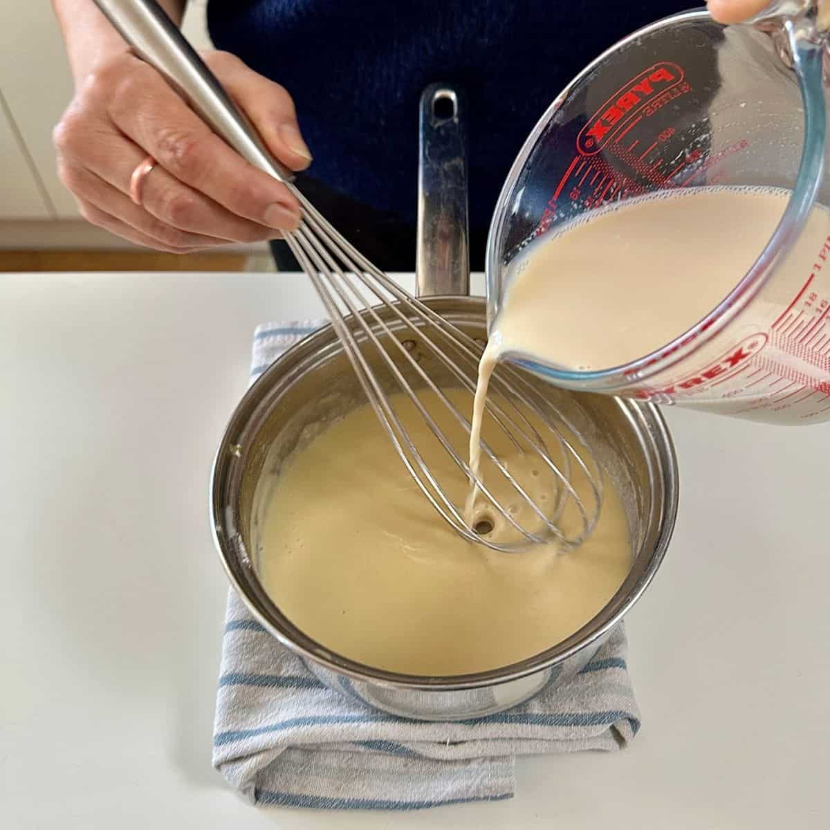 Pouring plant-based milk from a glass measuring jug into a steel saucepan containing a butter/flour paste, incorporating together with a whisk.