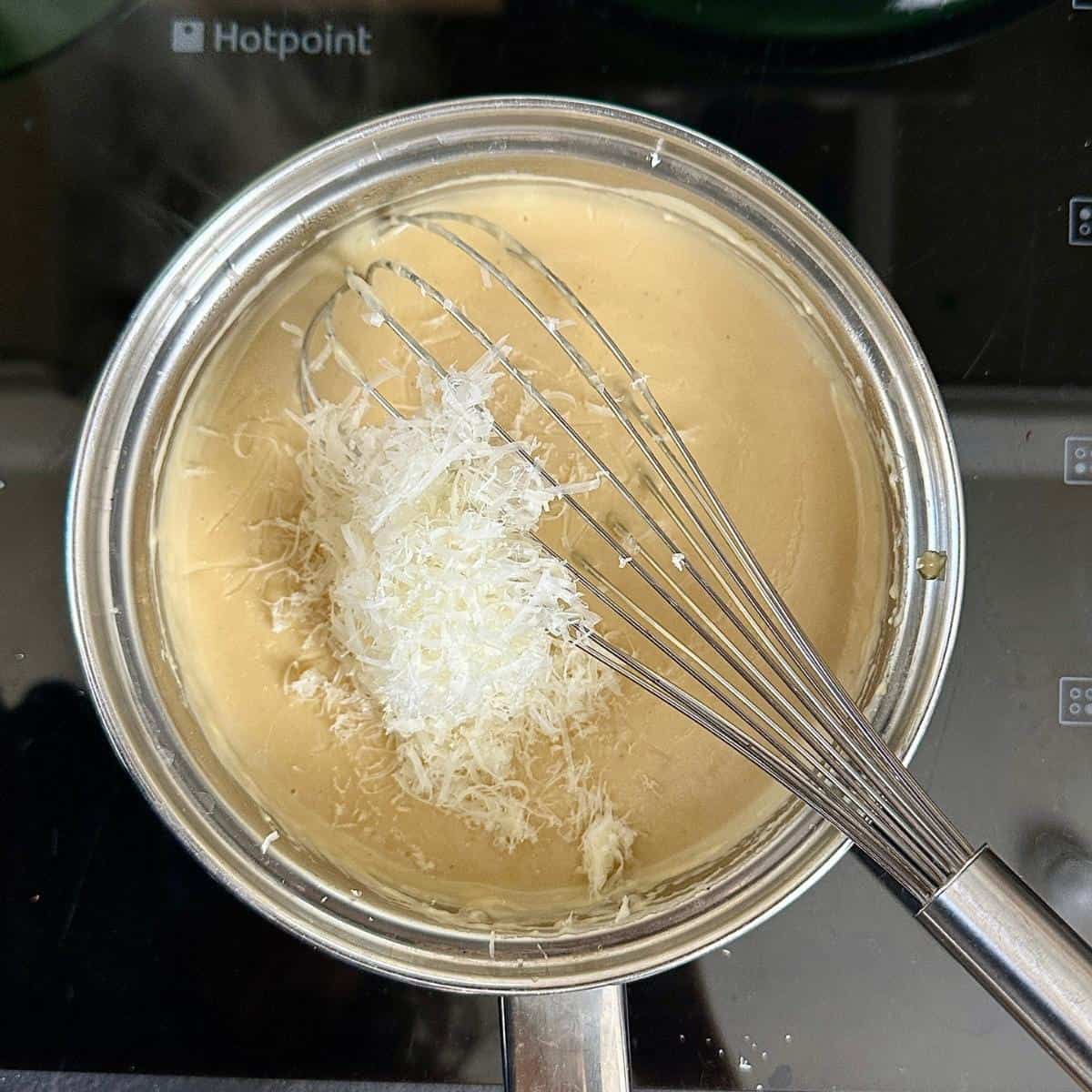 A steel saucepan containing a cheese sauce with grated vegan parmesan cheese on the surface ready to whisk into the sauce. A whisk rests in the sauce.