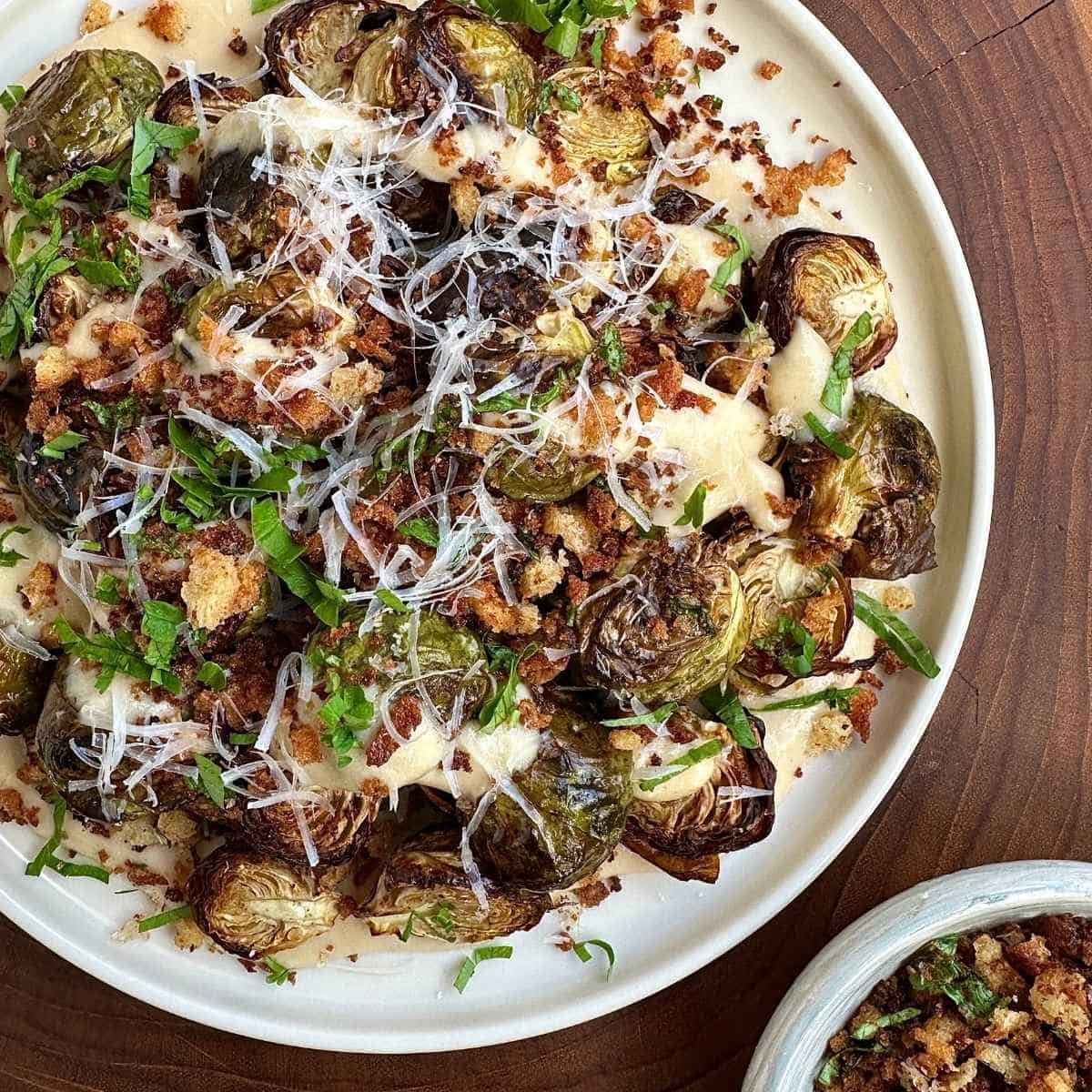 A white plate with crispy roasted Brussels sprouts in a vegan cheese sauce, sprinkled with grated vegan parmesan and parsley.