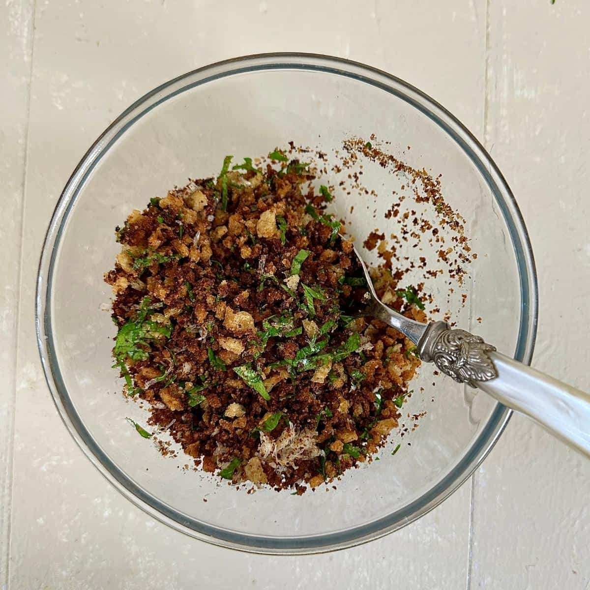 A glass bowl containing fried sourdough breadcrumbs with grated vegan parmesan cheese and parsley mixed into it.