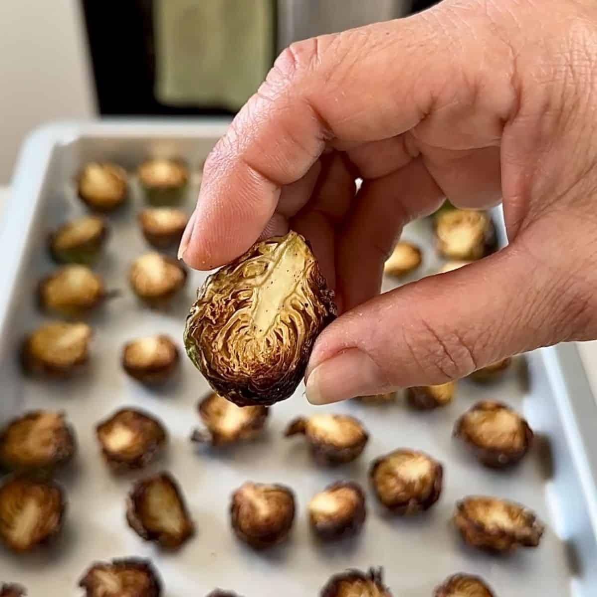 A hand holding a roast Brussels sprout close to the camera. A baking tray with more roasted Brussels sprouts sits in the background.