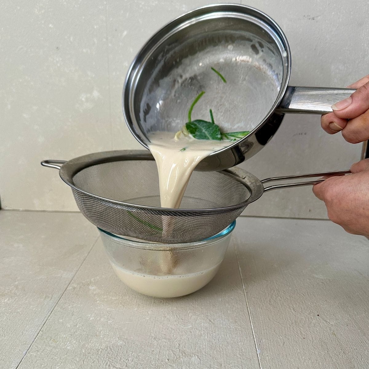 Pouring plant-based milk infused with onion, bay leaf, peppercorn and parsley from a steel saucepan, through a sieve into a glass bowl.