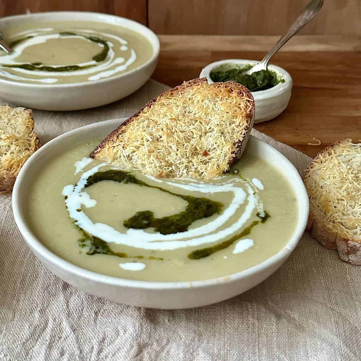 caramelised onion and leek soup in a beige ceramic bowl with a vegan cream and pesto garnish. A slice of cheesy bread sits in the soup.