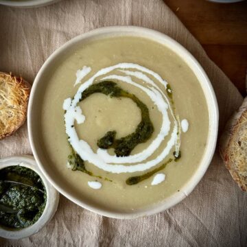 A beige ceramic bowl containing caramelised onion and leek soup, garnished with swirls of dairy-free cream and a green pesto.