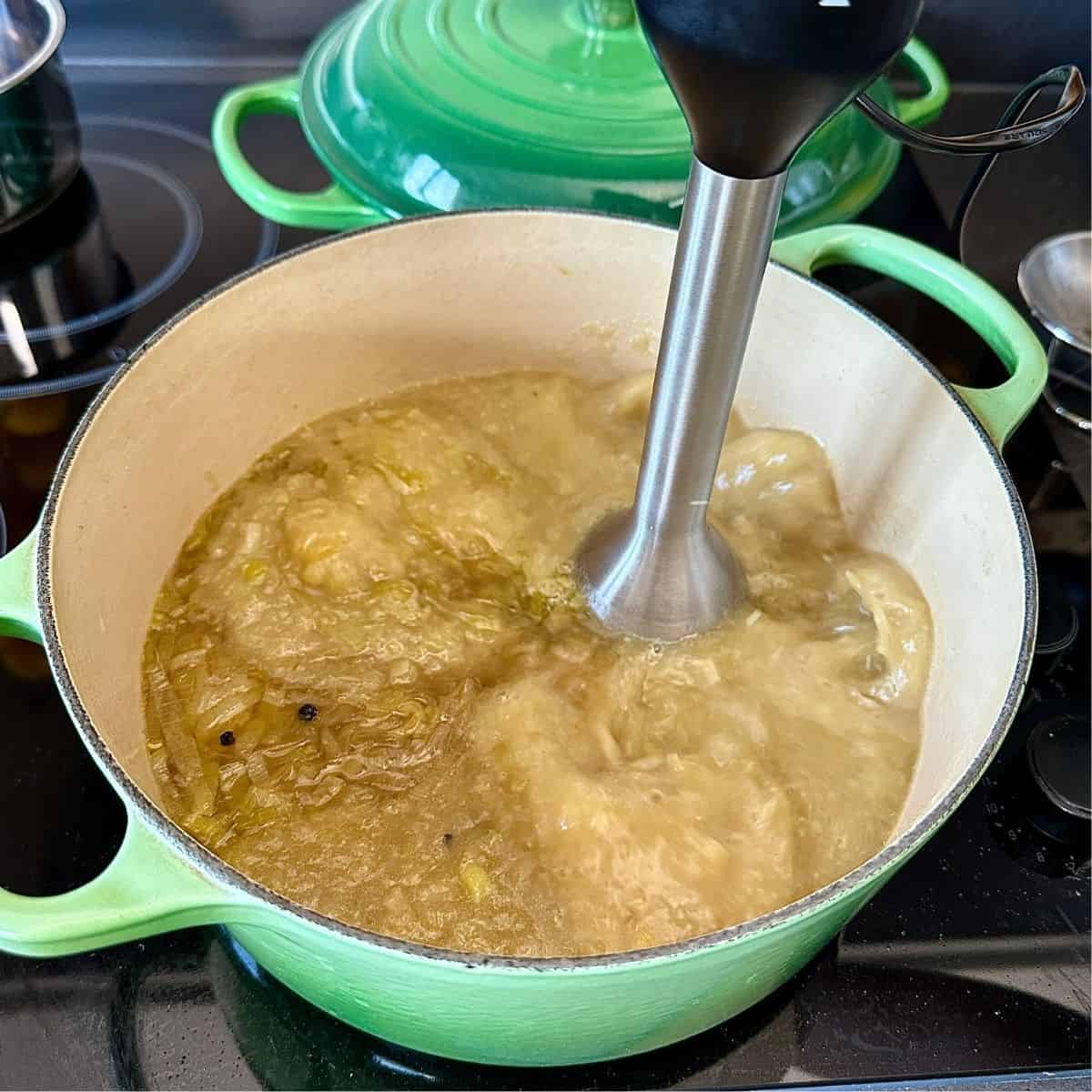 A large green pot containing onion and leek soup being pureed with an electric stick blender.