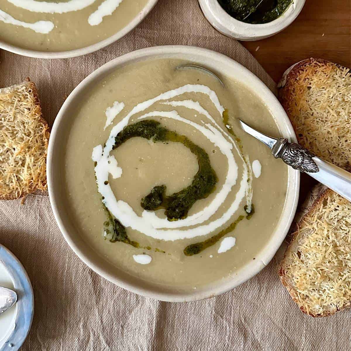 A beige ceramic bowl containing caramelised onion and leek soup, garnished with swirls of vegan cream and pesto.