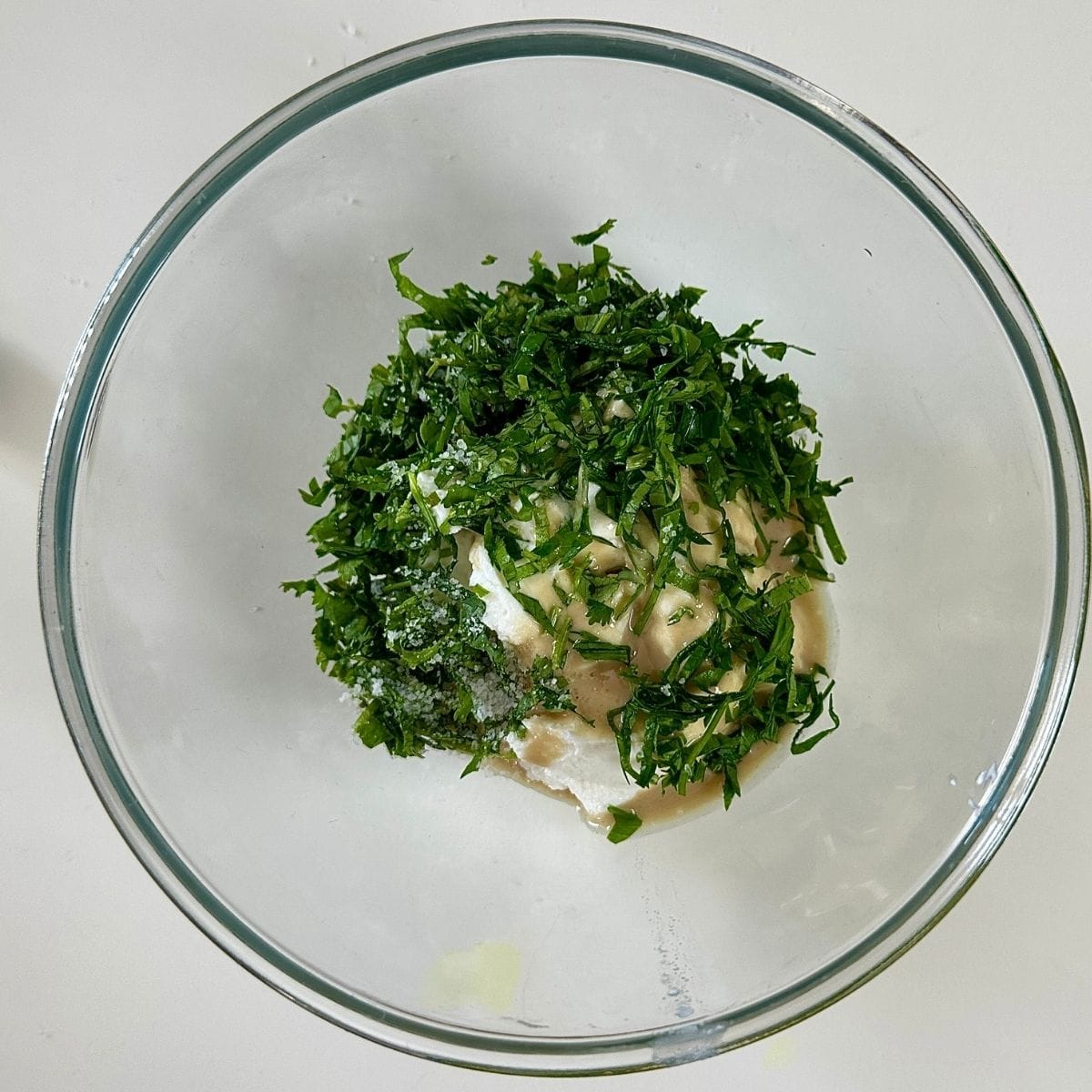 A glass bowl containing vegan yoghurt, tahini, chopped coriander and parsley and salt, ready to mix together to make a yoghurt tahini dip.